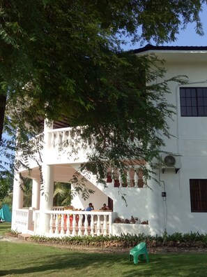 A white building with a balcony, a staircase, and a large tree in front.