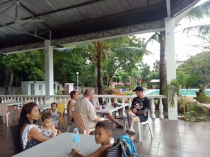An outdoor covered dining area with tables and chairs, surrounded by greenery and a pool.