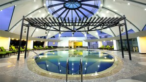 An indoor swimming pool with a central skylight, surrounded by lounge chairs and a covered seating area. - BCDM Rajendrapur (Pirozali)