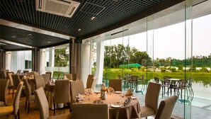 A restaurant with tables set for dining, large windows offering a view of a golf course, and a modern ceiling design. - BCDM Rajendrapur (Pirozali)