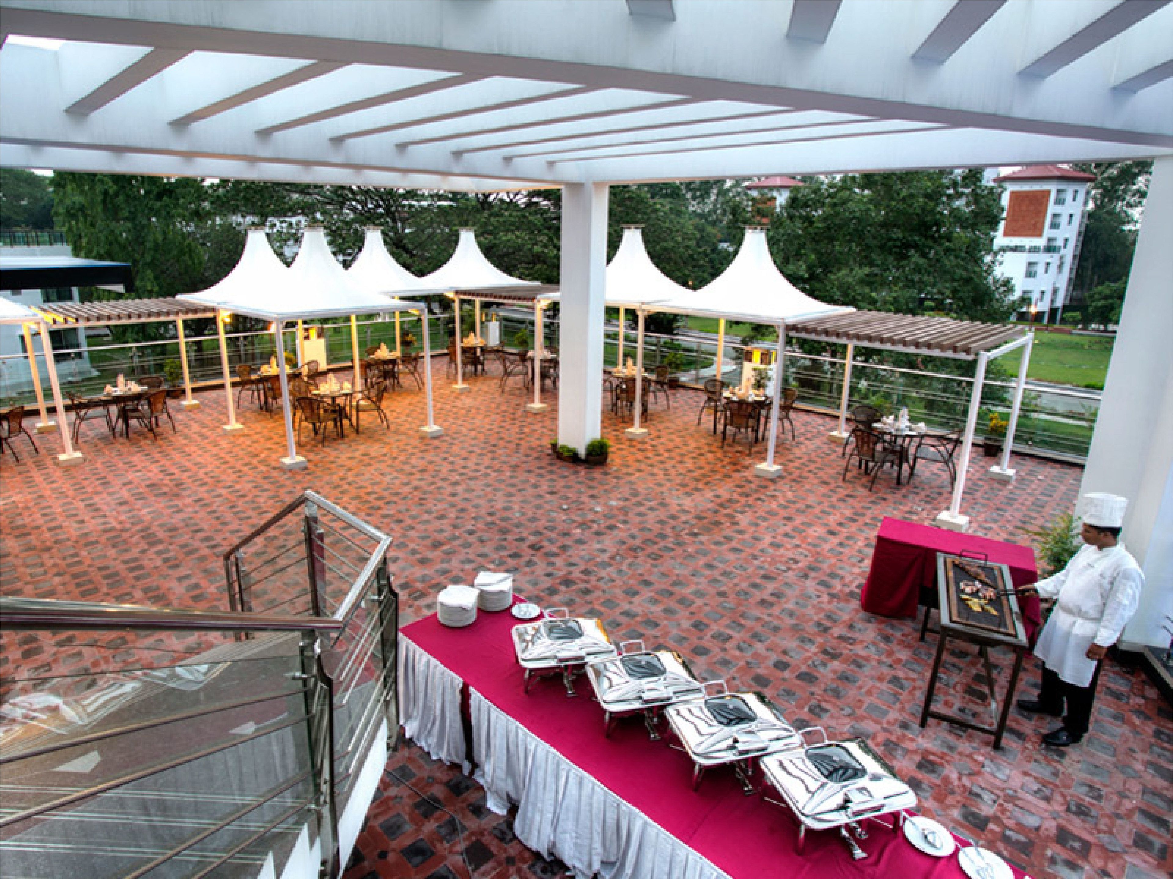 An outdoor dining area with tables and chairs under white canopies, a chef at work, and a view of greenery and buildings in the background.