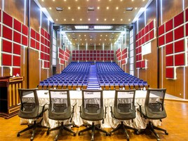 Un salón de conferencias con filas de sillas azules, un atril y paneles rojos en las paredes.