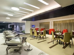 A modern dining area with multiple tables and chairs, a flat-screen TV mounted on the wall, and a large window with curtains.