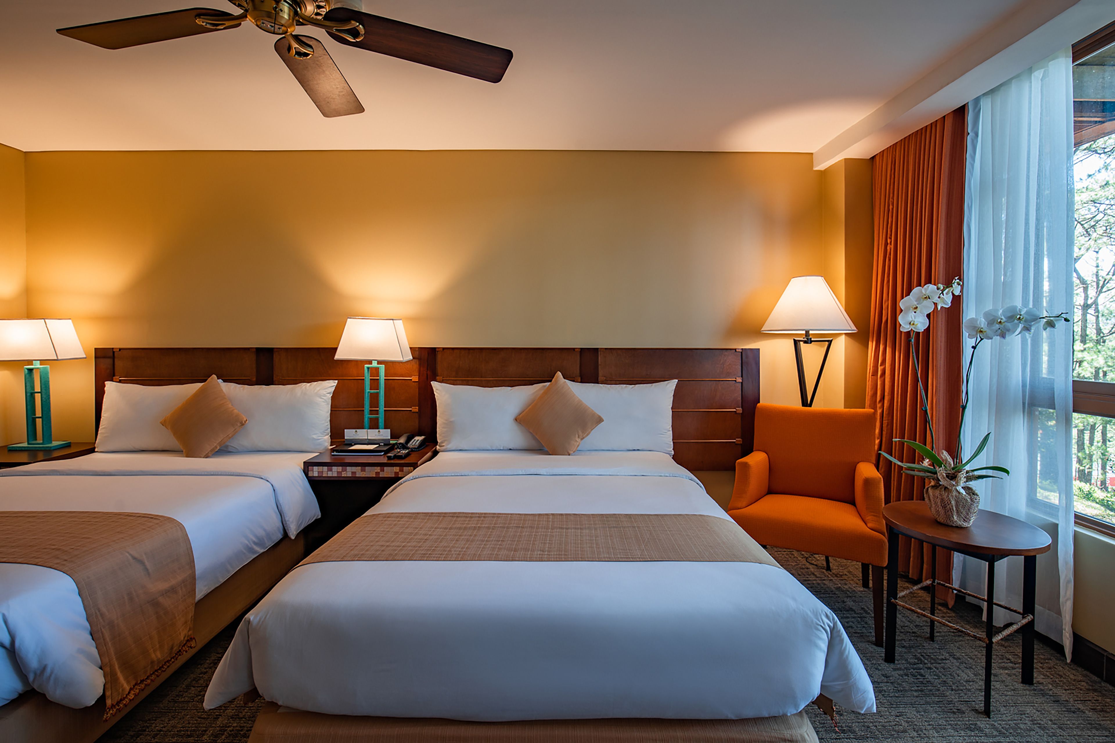 A hotel room with two beds, a ceiling fan, a chair, a small table with a plant, and a window with curtains.