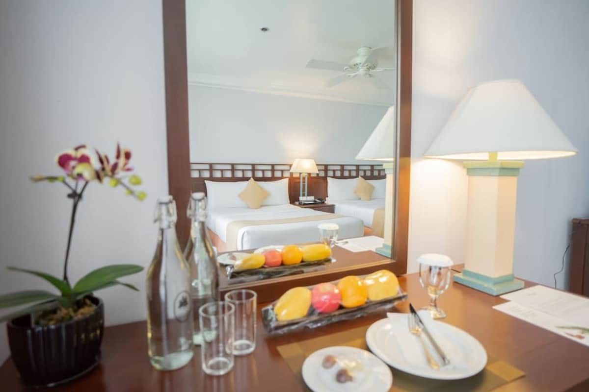 Une chambre d’hôtel avec un lit, une table de chevet avec une lampe, une assiette de fruits, un vase de fleurs et un miroir qui reflète la pièce.