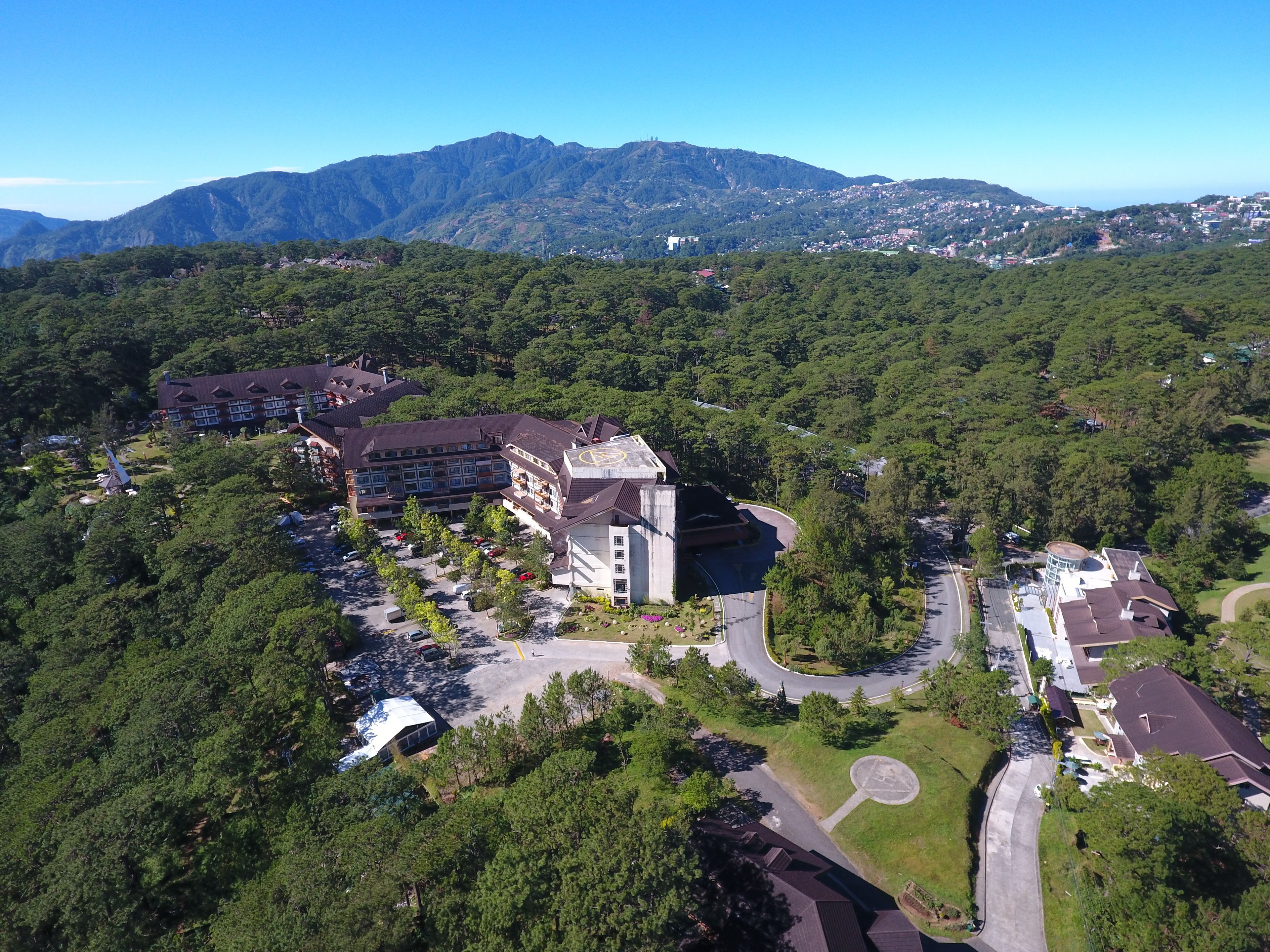 A hotel with a central building surrounded by lush greenery, a parking area, and a view of distant mountains.