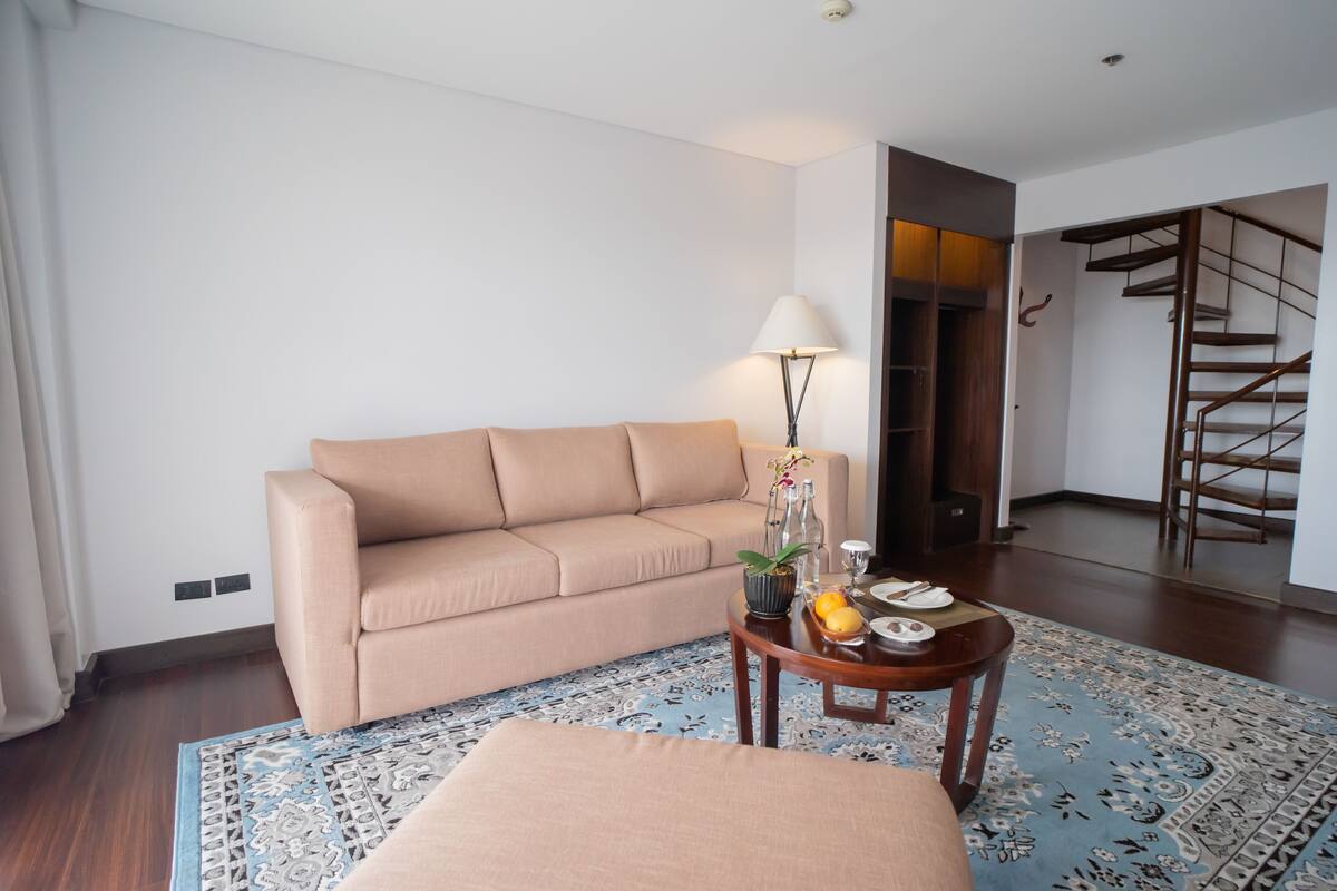 A living room with a beige sofa, a round wooden coffee table with a lamp, and a patterned rug.