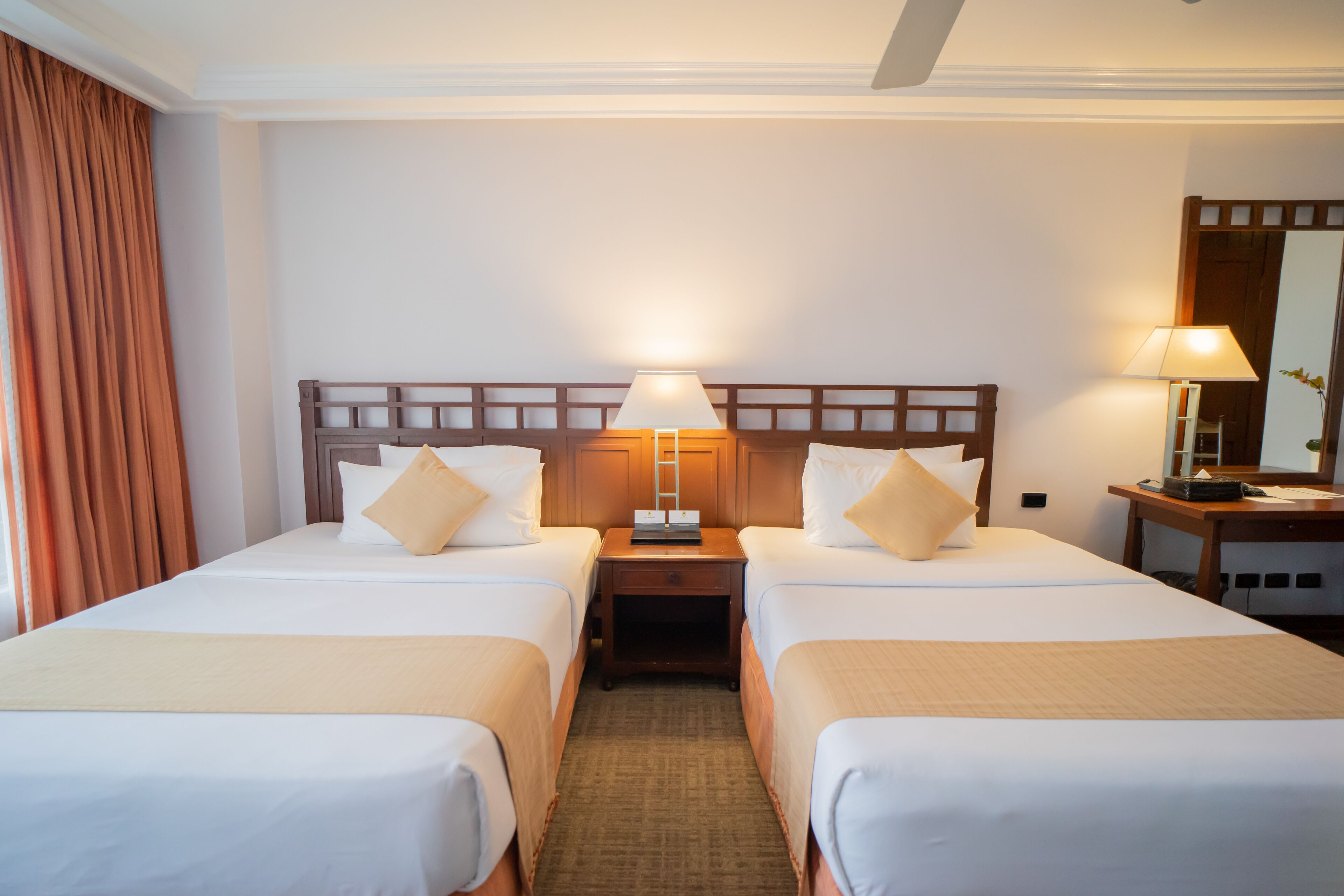 Habitación de hotel con dos camas, una mesita de noche con lámpara, un espejo y una ventana con cortinas.