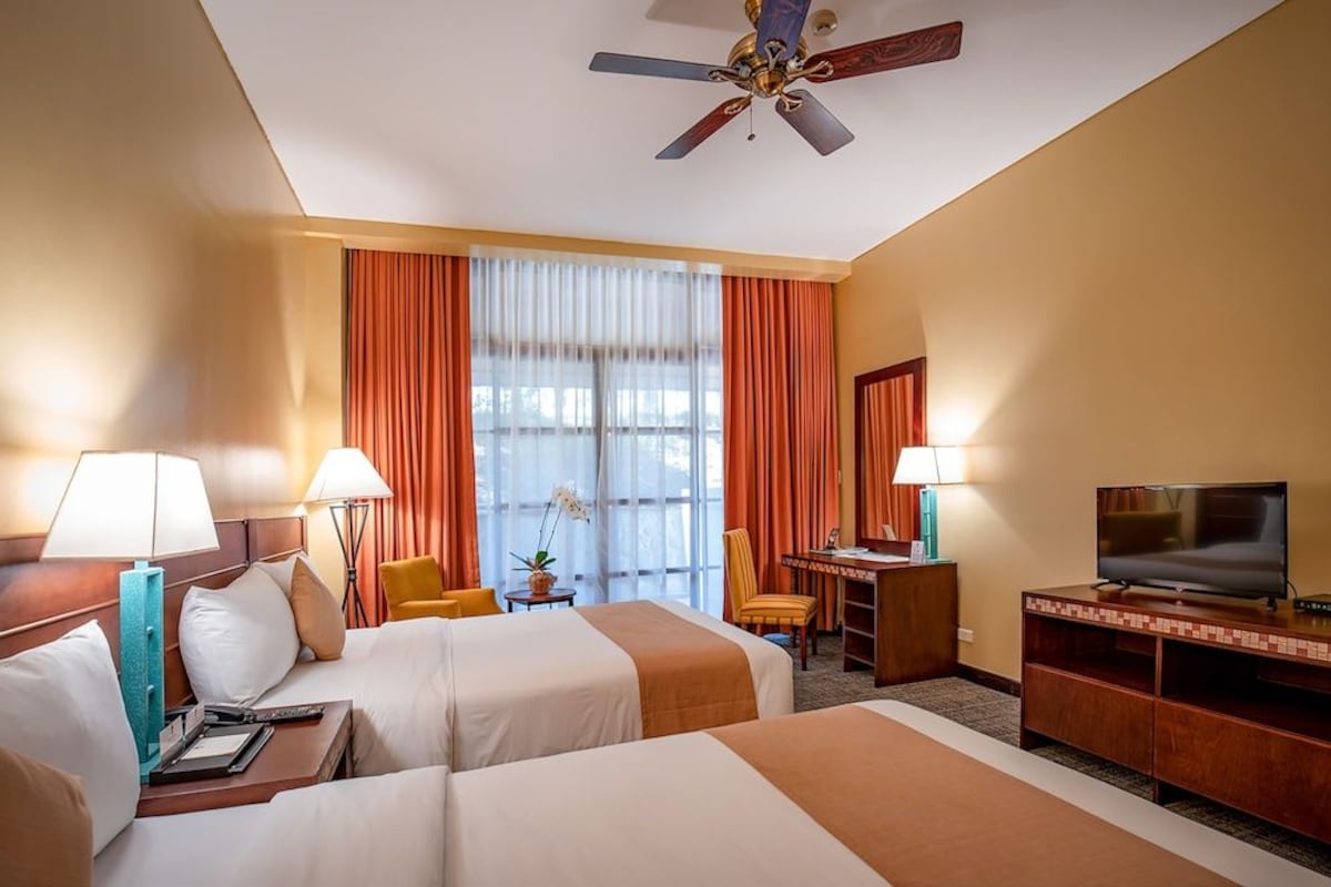 A hotel room with two beds, a desk, a television, and a ceiling fan.