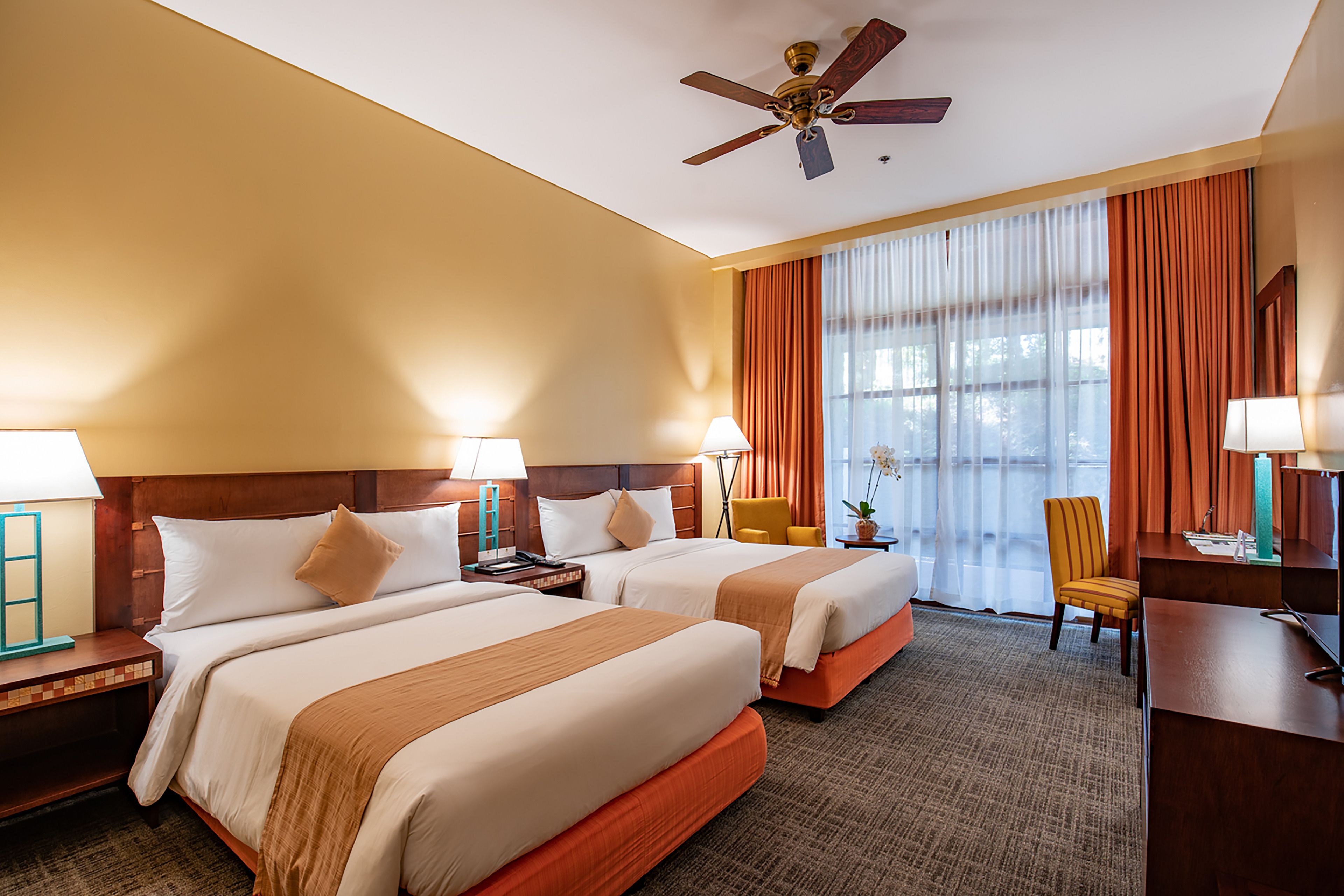 A hotel room with two beds, a desk, a chair, a ceiling fan, and a large window with curtains.