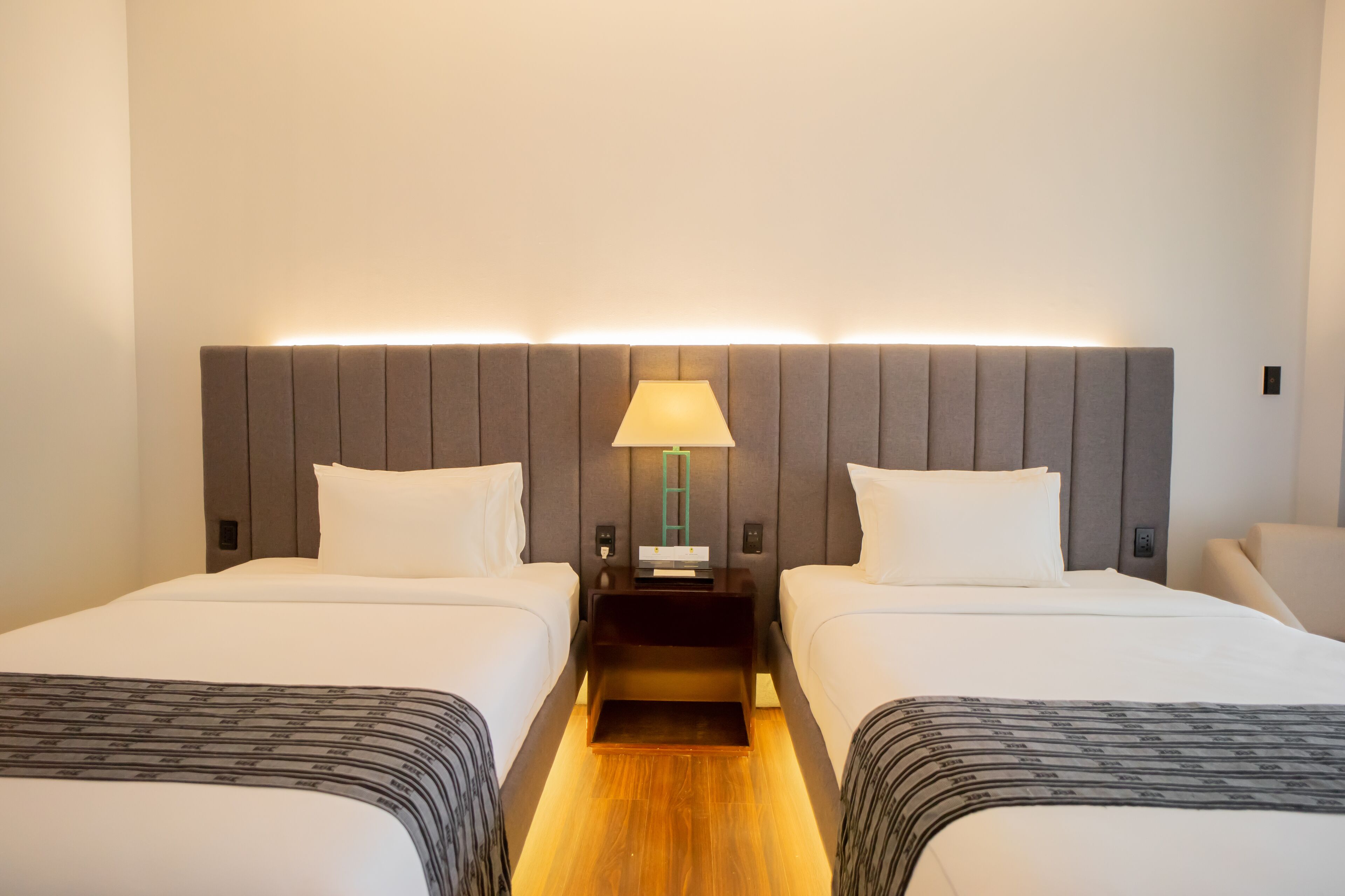 A hotel room with two beds, a nightstand with a lamp, and a wooden floor.