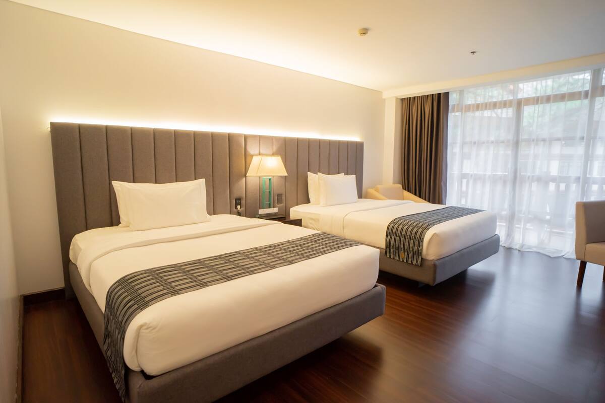 A hotel room with two beds, a nightstand with a lamp, a chair, and a large window with curtains.