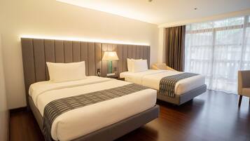 Habitación de hotel con dos camas, una mesita de noche con lámpara, una silla y un ventanal con cortinas.