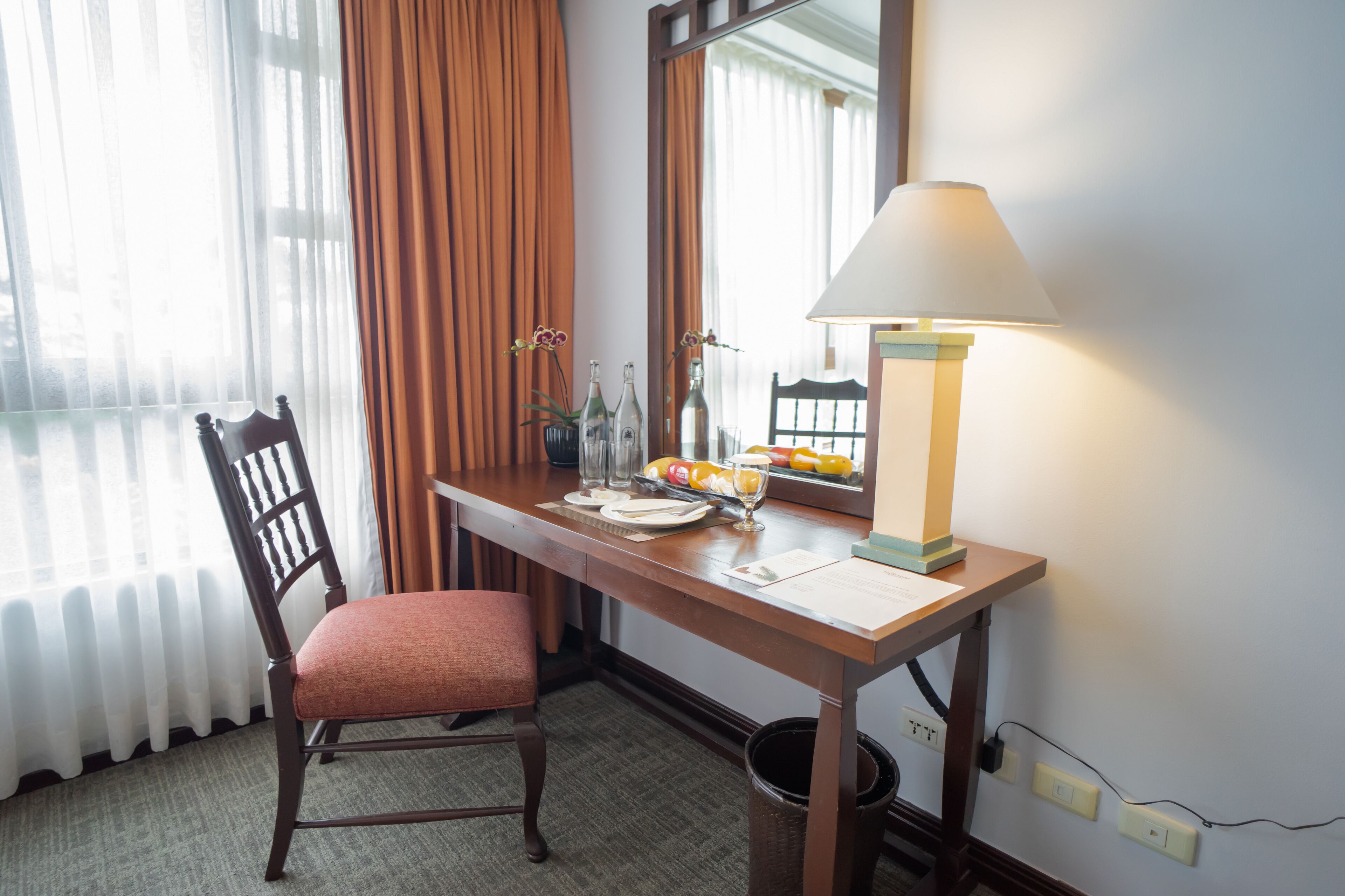 A hotel room with a wooden desk, a chair, a lamp, and a window with curtains.
