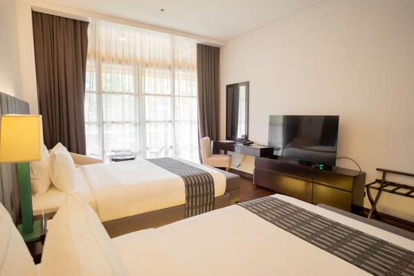 Habitación de hotel con dos camas, un escritorio, un televisor y un ventanal grande con cortinas.