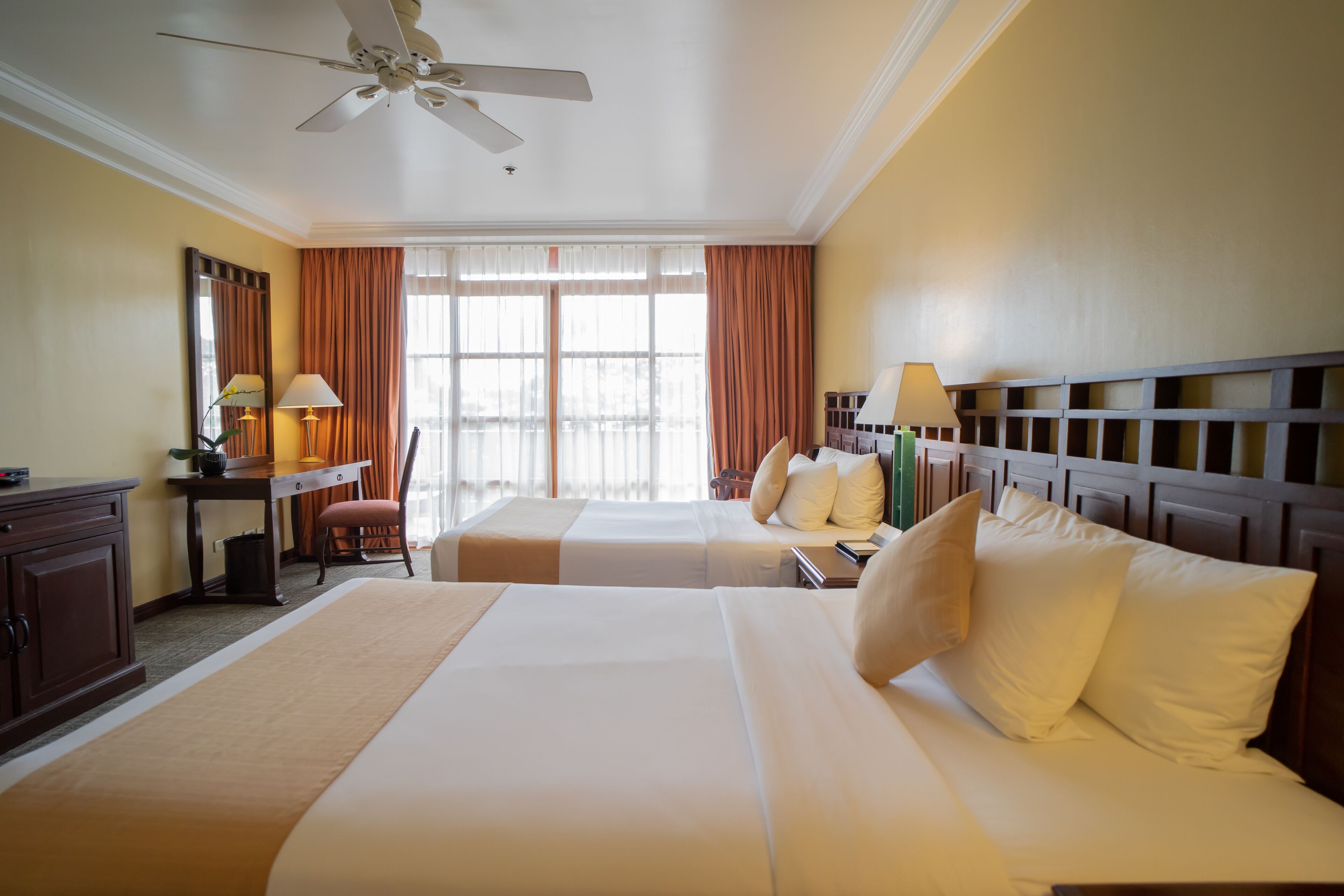 A hotel room with two beds, a desk with a chair, a ceiling fan, and large windows with curtains.