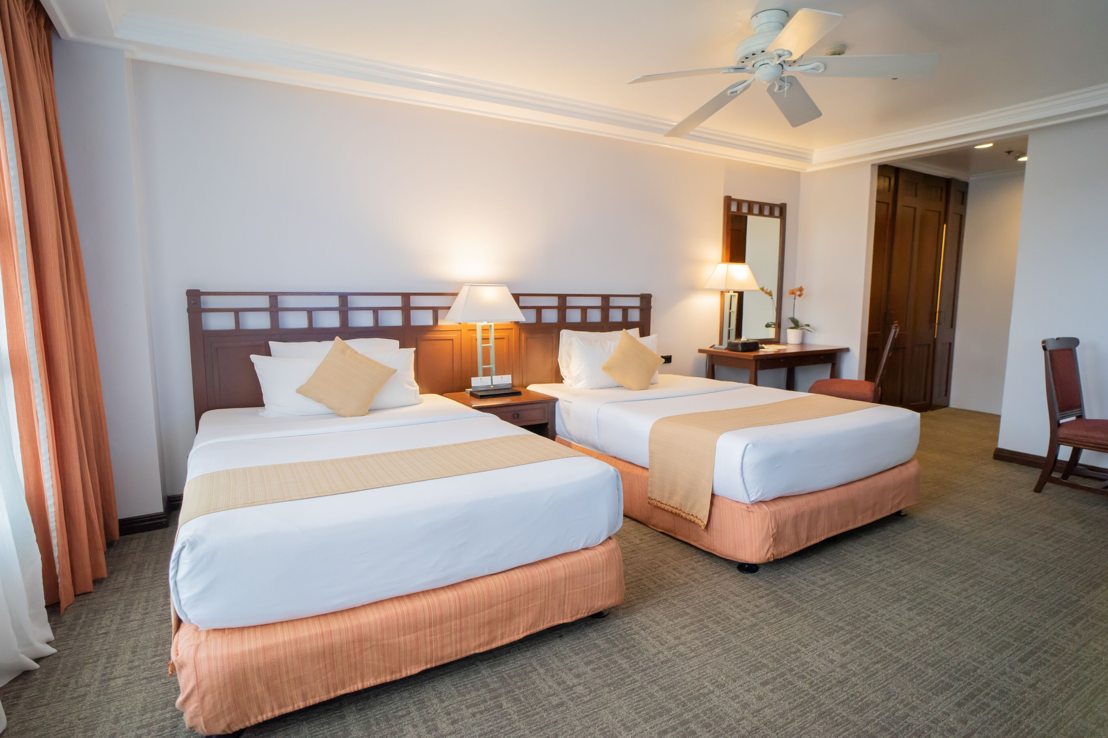 A hotel room with two beds, a desk, a chair, a ceiling fan, and a mirror.