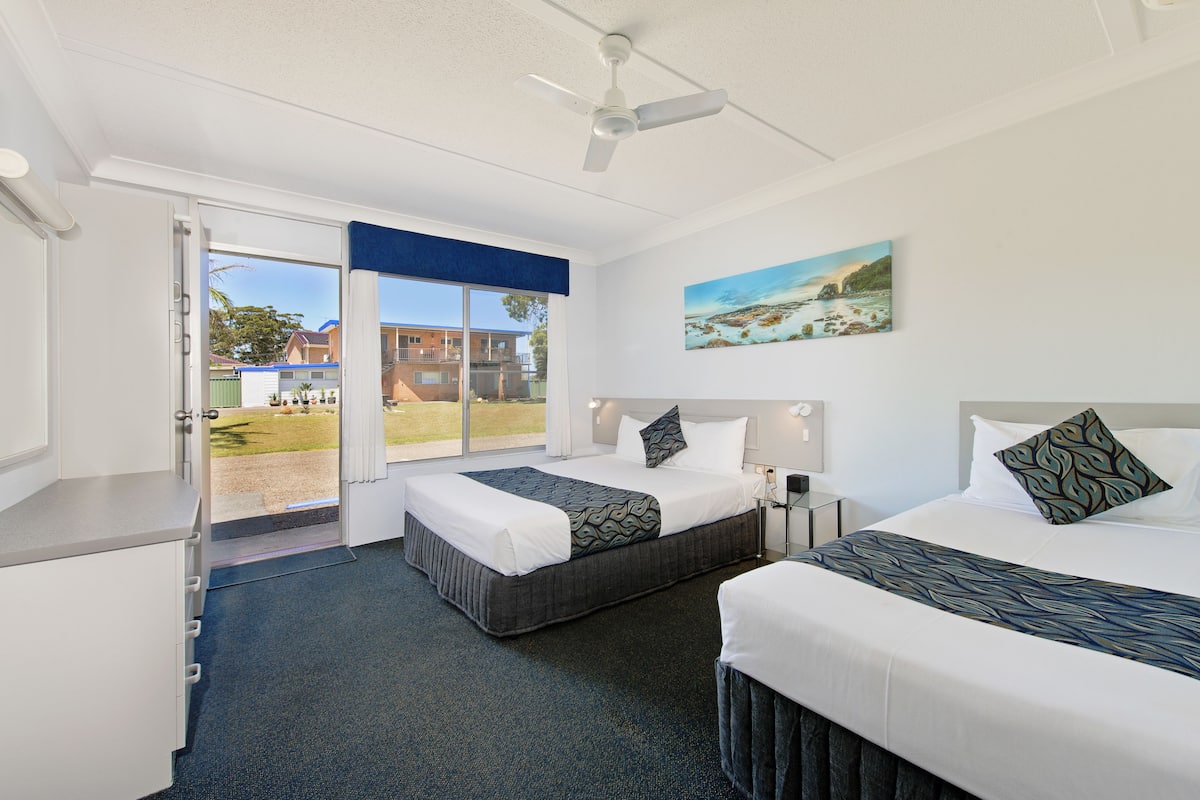 A hotel room with two beds, a ceiling fan, a large window with a view of buildings, and a painting on the wall.