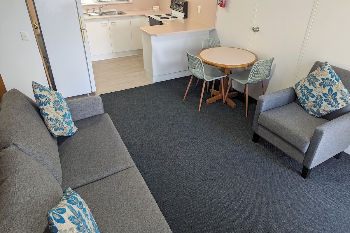 A compact living space with a kitchenette, grey sofas with patterned cushions, a round dining table with chairs, and a dark grey carpet.