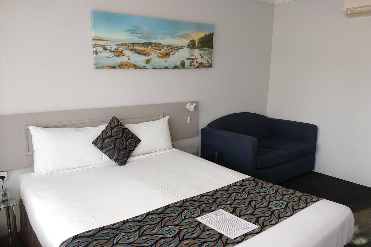 A hotel room with a bed, a chair, a painting on the wall, and a bedside table.