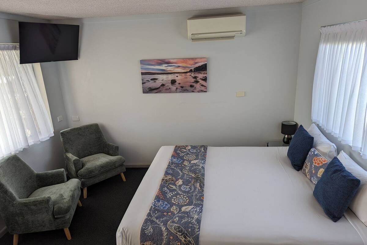 A hotel room with a bed, two armchairs, a flat-screen TV, an air conditioner, a painting, and a window with curtains.