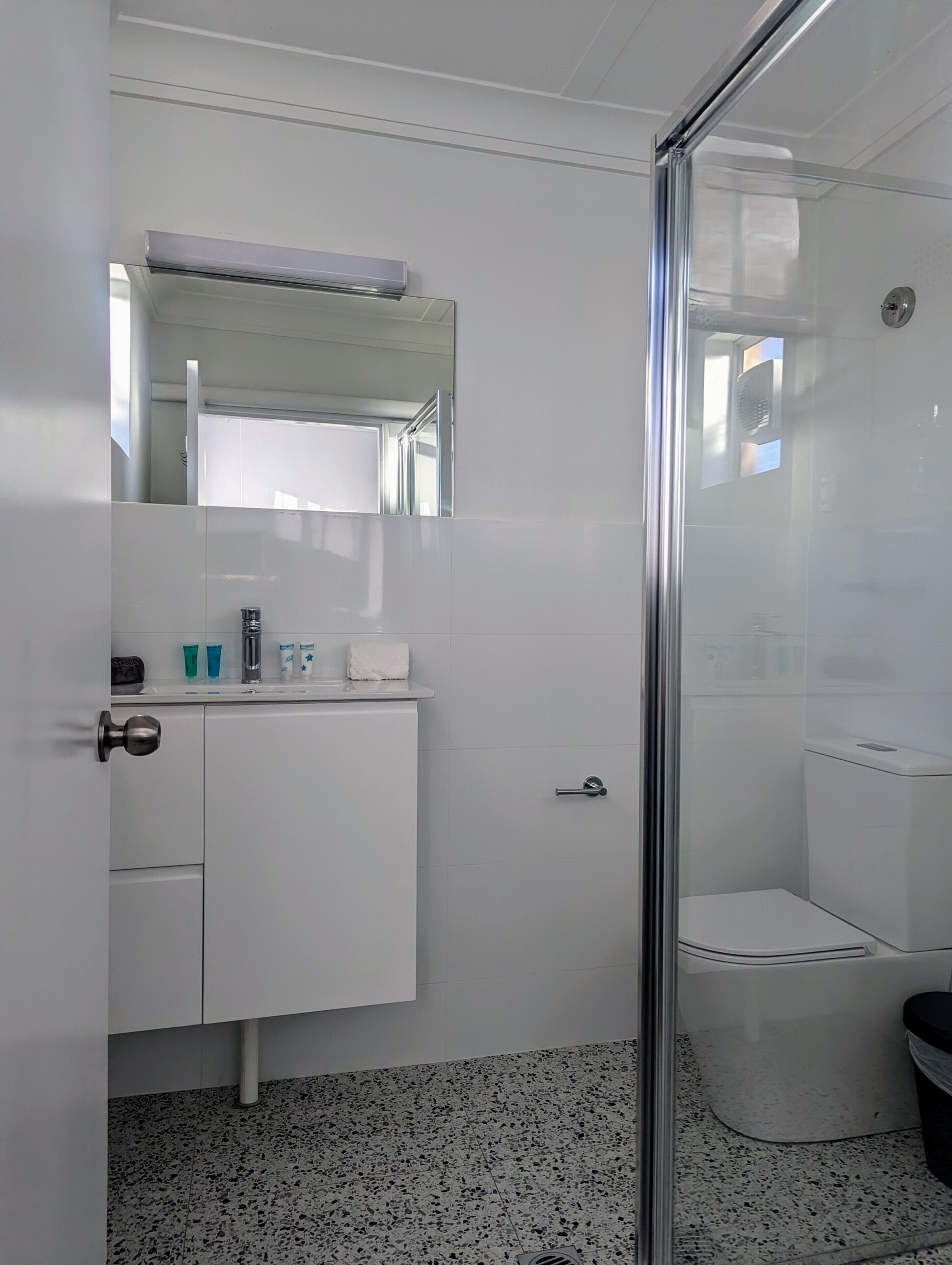 Uma casa de banho moderna com cabine de duche em vidro, uma sanita e um lavatório branco com artigos de higiene pessoal.