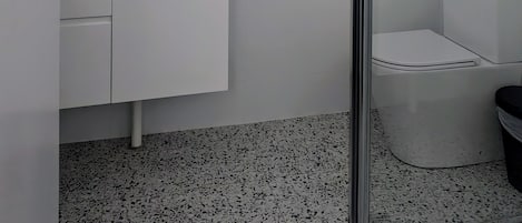 Uma casa de banho moderna com cabine de duche em vidro, uma sanita e um lavatório branco com artigos de higiene pessoal.