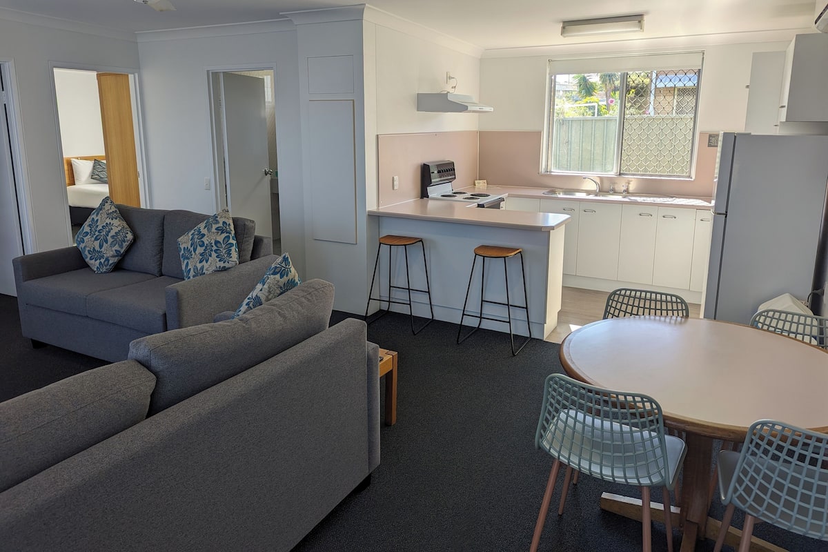 A modern living room with a grey sofa, two bar stools, a dining table with chairs, and a kitchen area with a window.
