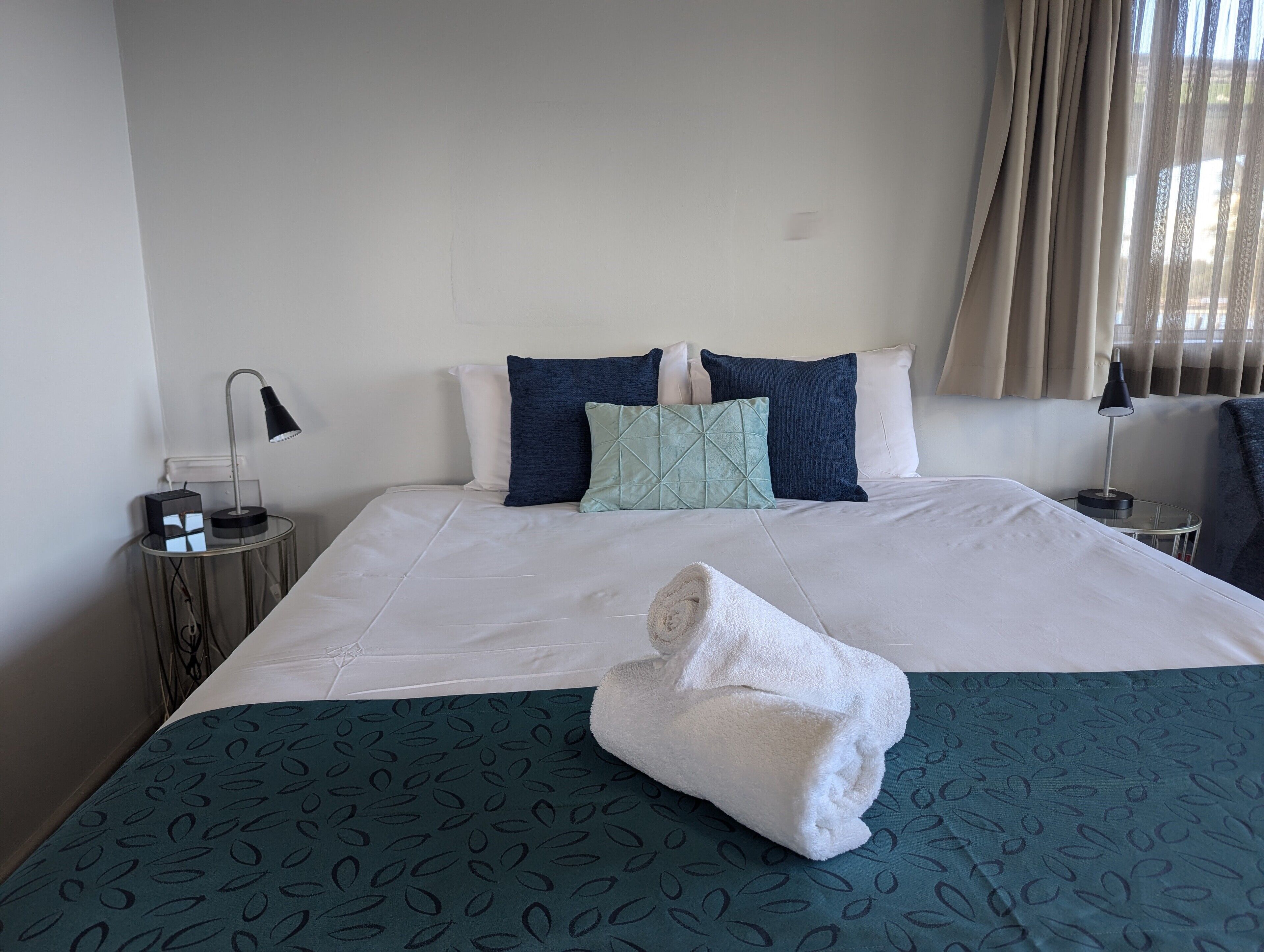 Une chambre d’hôtel avec un lit bien fait, une lampe de chevet, une petite table avec une lampe et une fenêtre avec des rideaux.