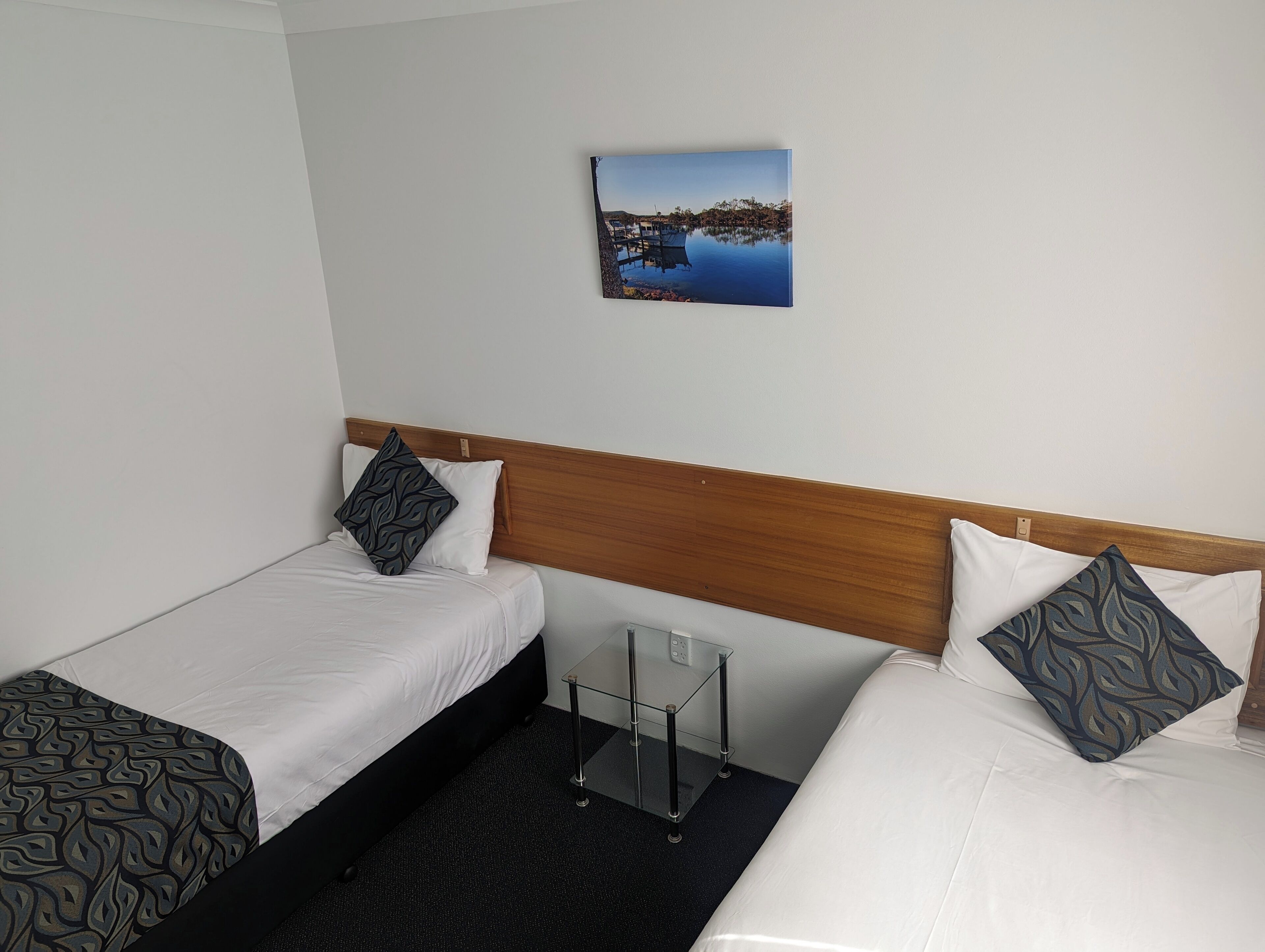 A hotel room with two single beds, a wooden headboard, a small bedside table, and a framed picture on the wall.