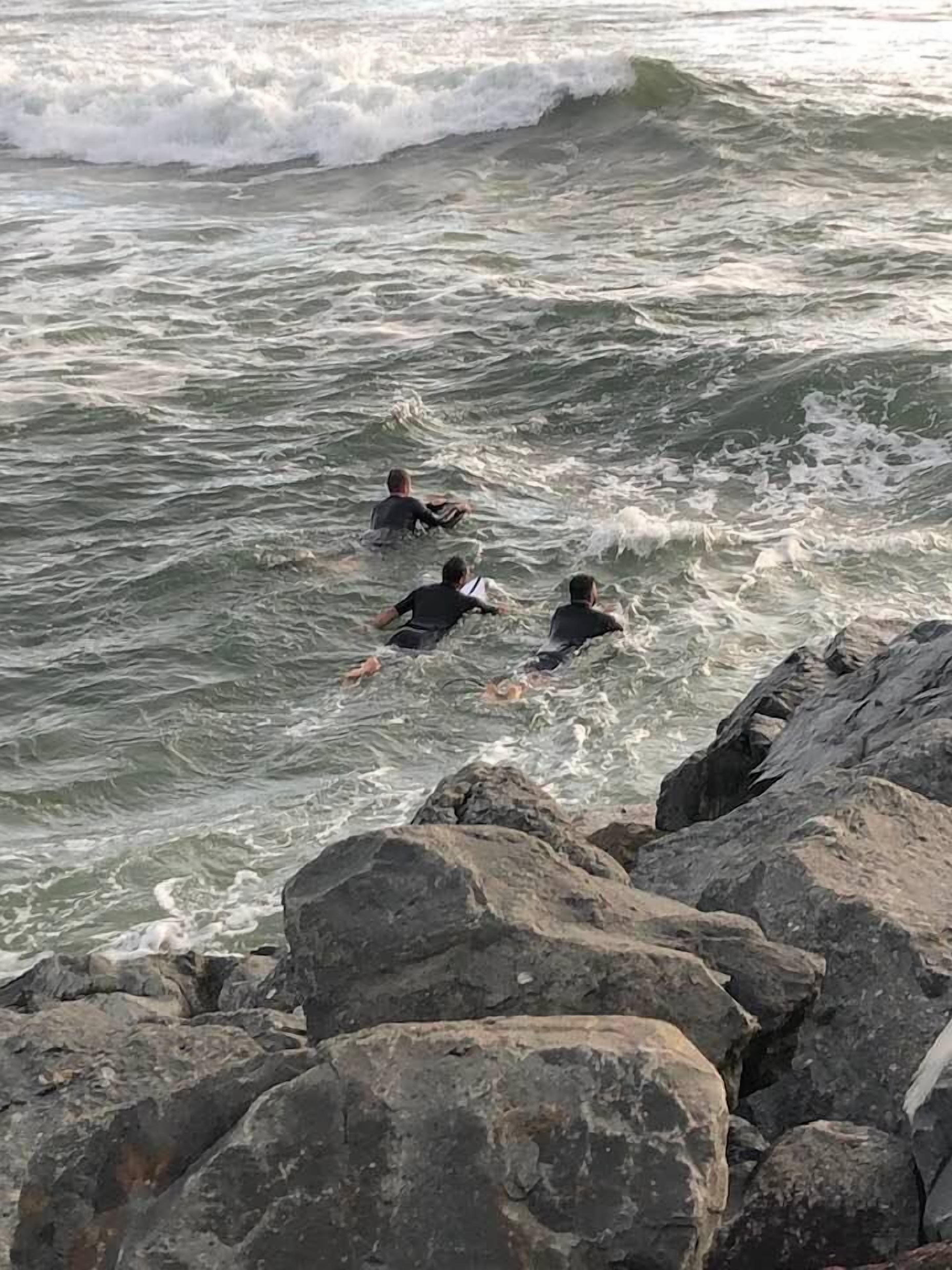 Tres personas nadando en aguas oceánicas agitadas cerca de grandes rocas.