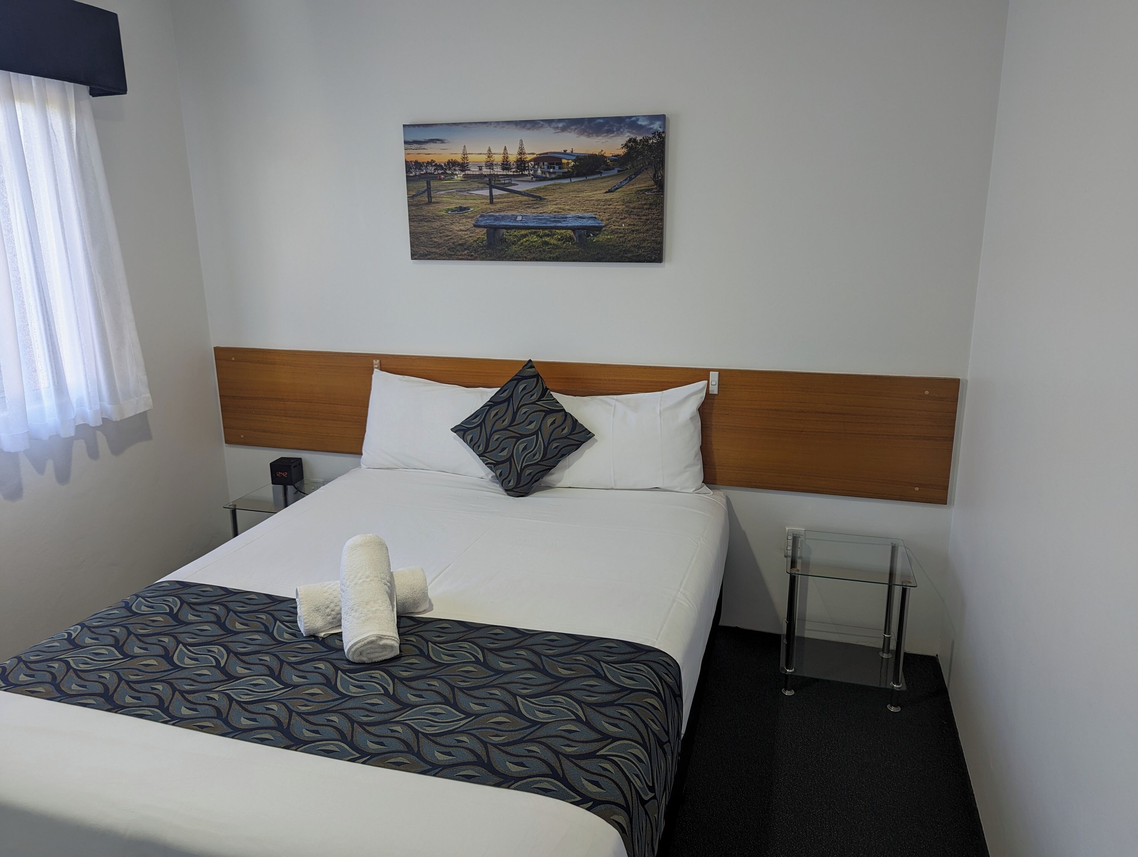 Habitación de hotel con cama, cabecero, mesita de noche, un cuadro colgado en la pared y una ventana con cortinas.