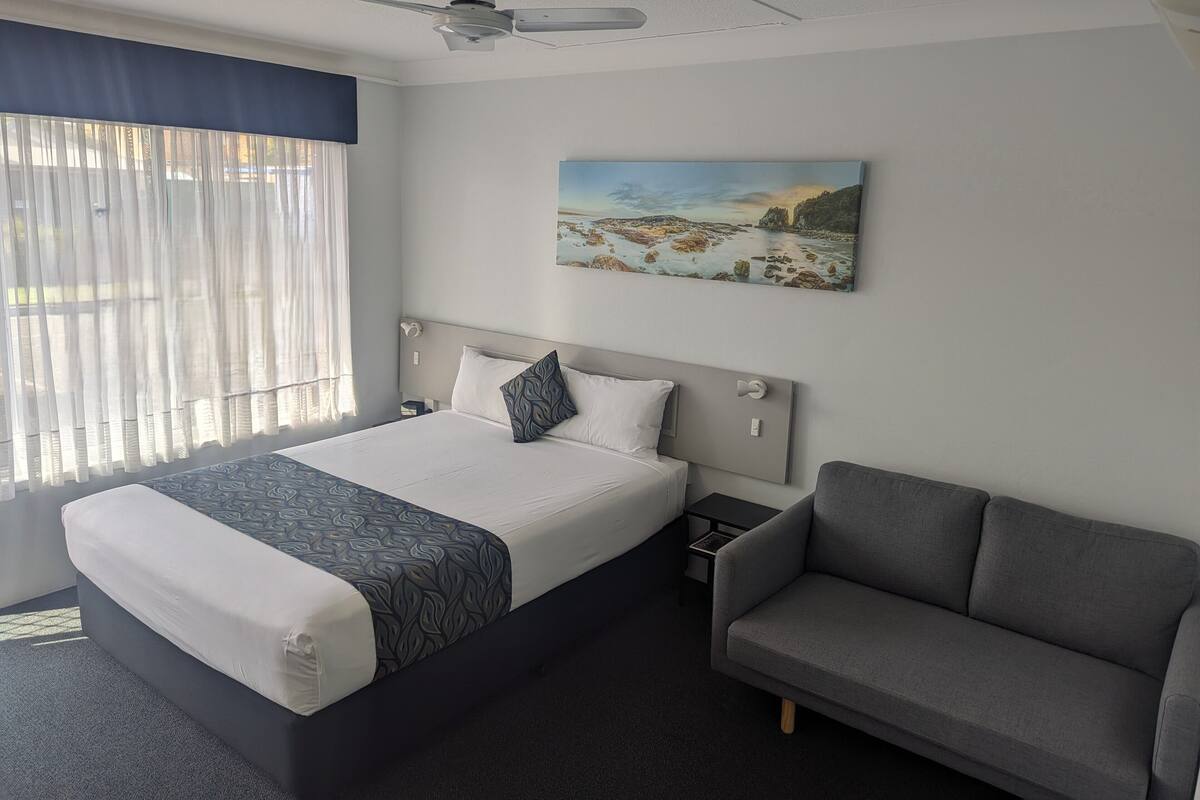 A hotel room with a bed, a gray sofa, a painting on the wall, and a ceiling fan.