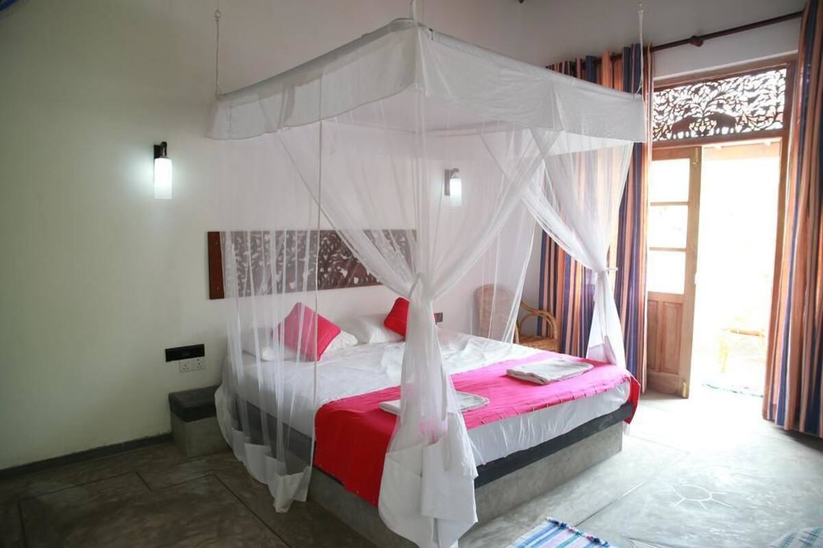 Un dormitorio con una cama bajo un dosel blanco, una colcha roja y blanca, y vistas al exterior a través de una ventana con cortinas.