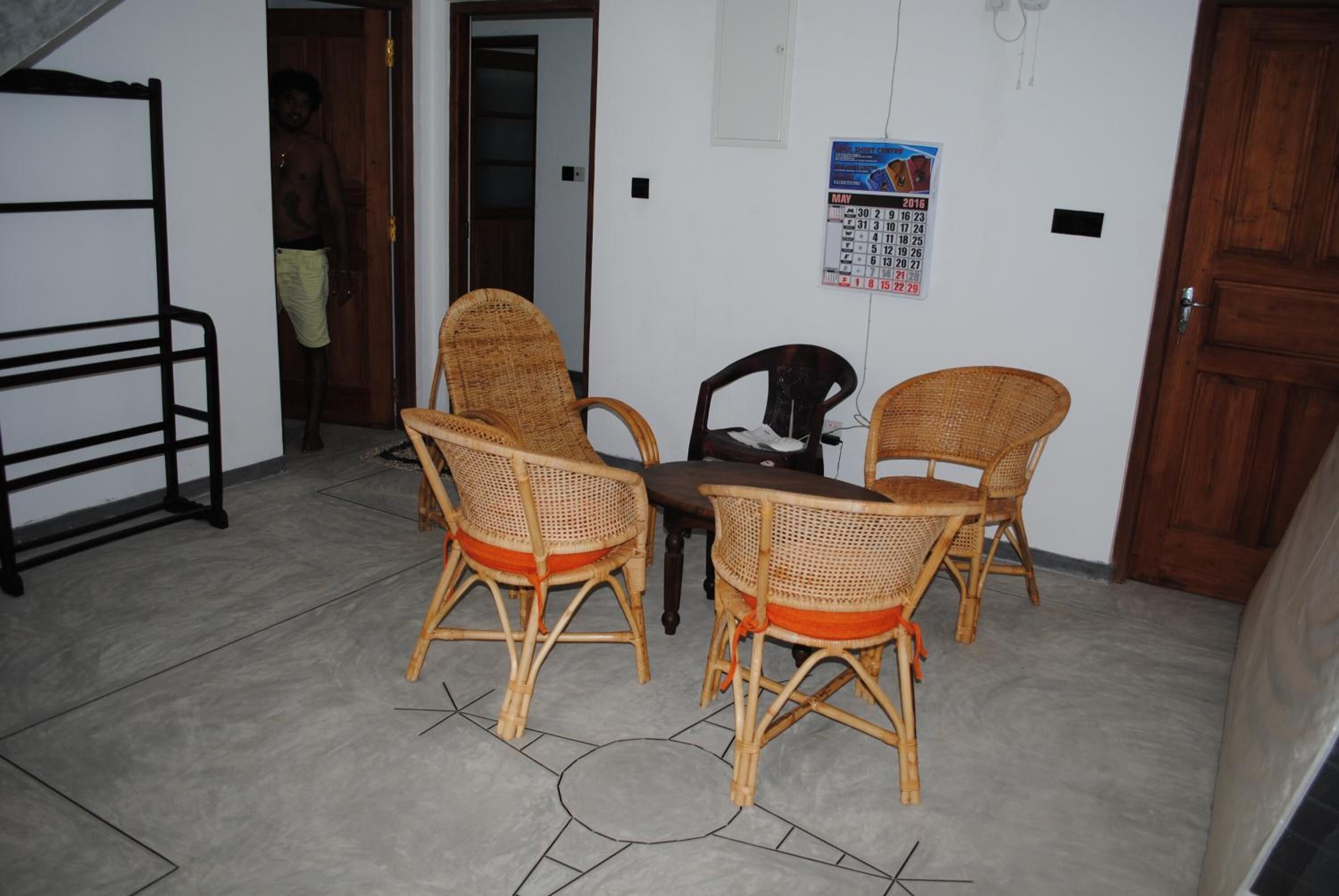 Une pièce avec une table et quatre chaises en osier, un calendrier accroché au mur et une personne se tenant dans l’embrasure de la porte.