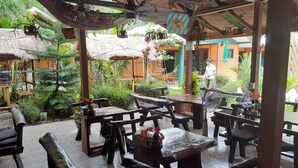 An outdoor patio area with wooden furniture, potted plants, and a thatched roof structure. - New Paradise Home Resort (Rayong)