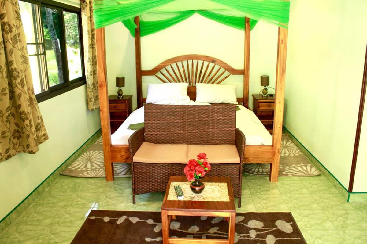 Une chambre avec un lit à baldaquin, une petite table sur laquelle est posé un vase de fleurs et une fenêtre avec des rideaux.