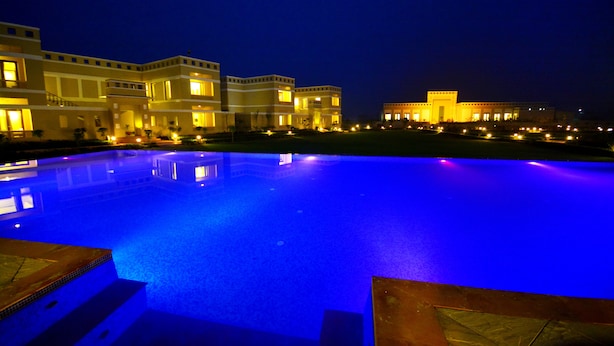 A nighttime view of a hotel with a swimming pool, illuminated buildings, and a clear sky. - Achrol Niwas A TreeHouse Hotel (Amer)