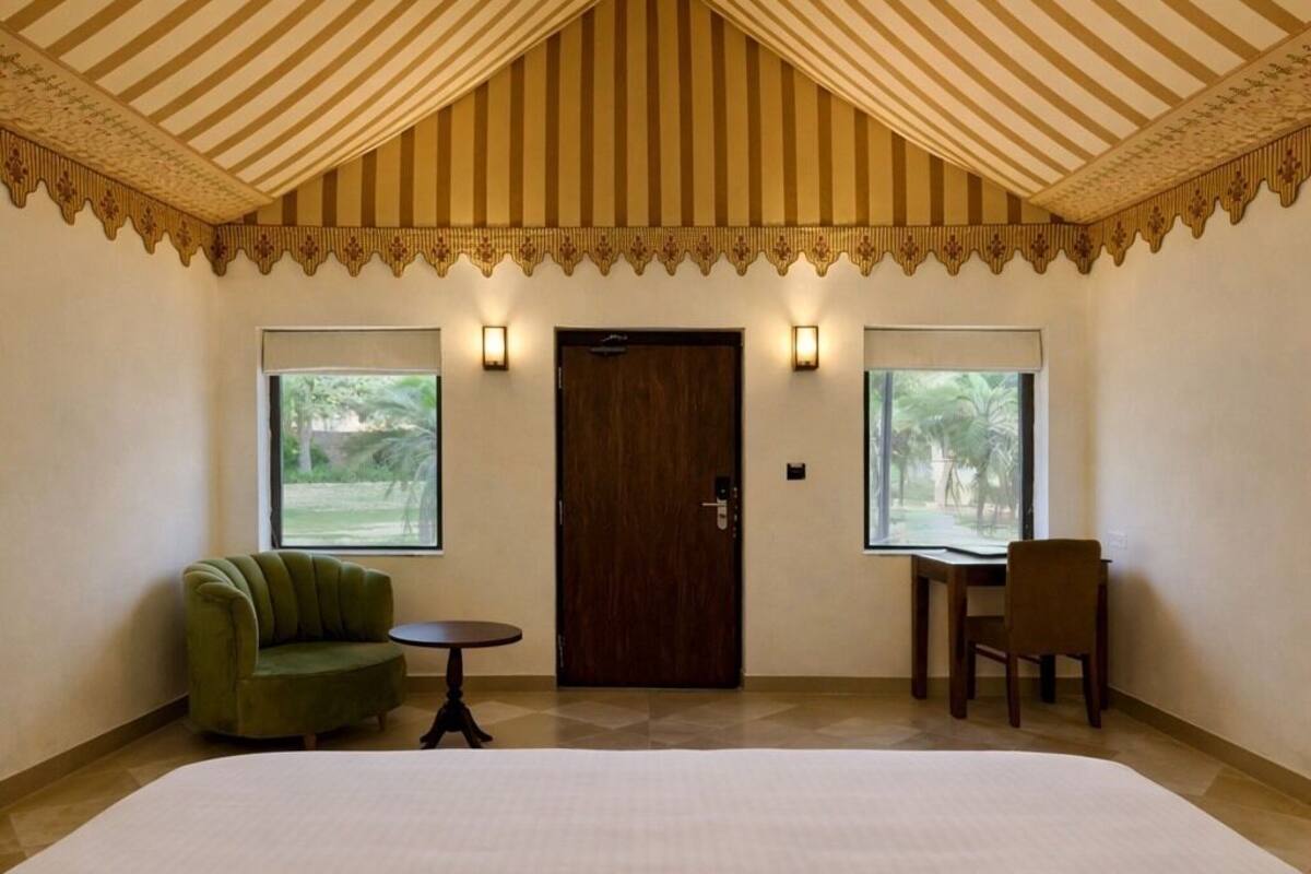 Quarto de hotel com uma cama grande, duas cadeiras, uma mesinha e duas janelas com vista para áreas verdes.
