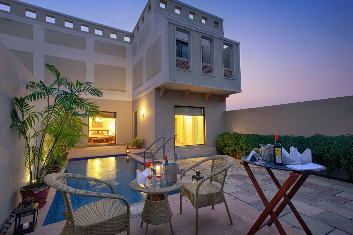Uma área de piscina externa com cadeiras de vime, uma mesa com bebidas e um edifício com varanda.