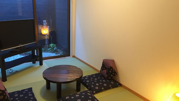 A room with tatami flooring, a low table, cushions, a television, and a view of a garden with a water feature.