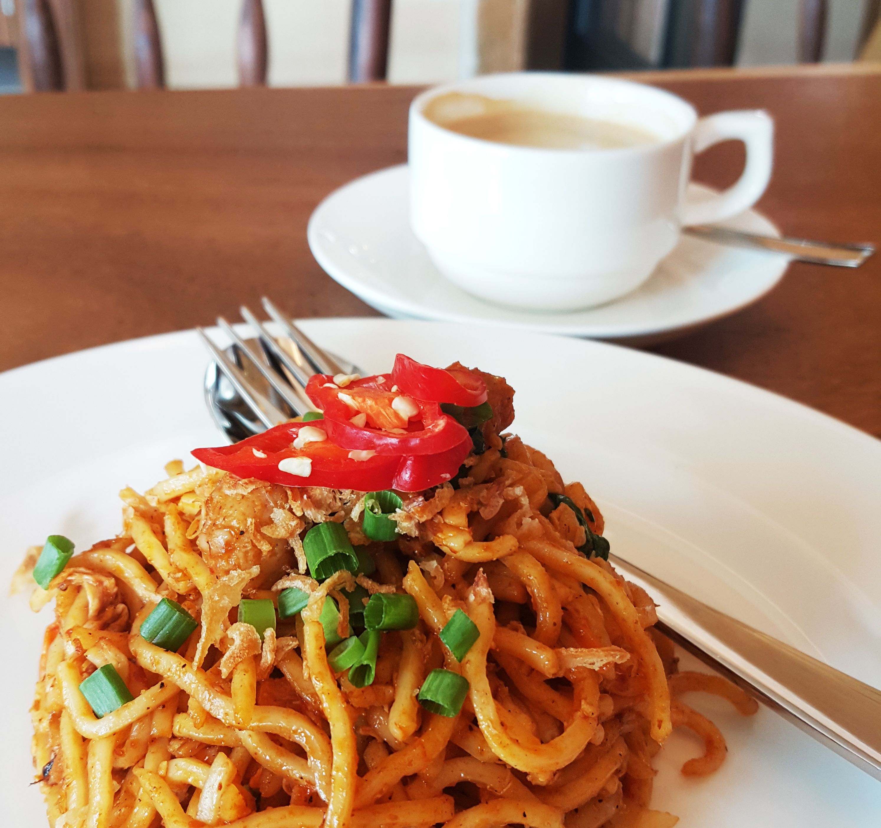 A plate of noodles garnished with red chili and green onions, accompanied by a cup of coffee on a saucer.