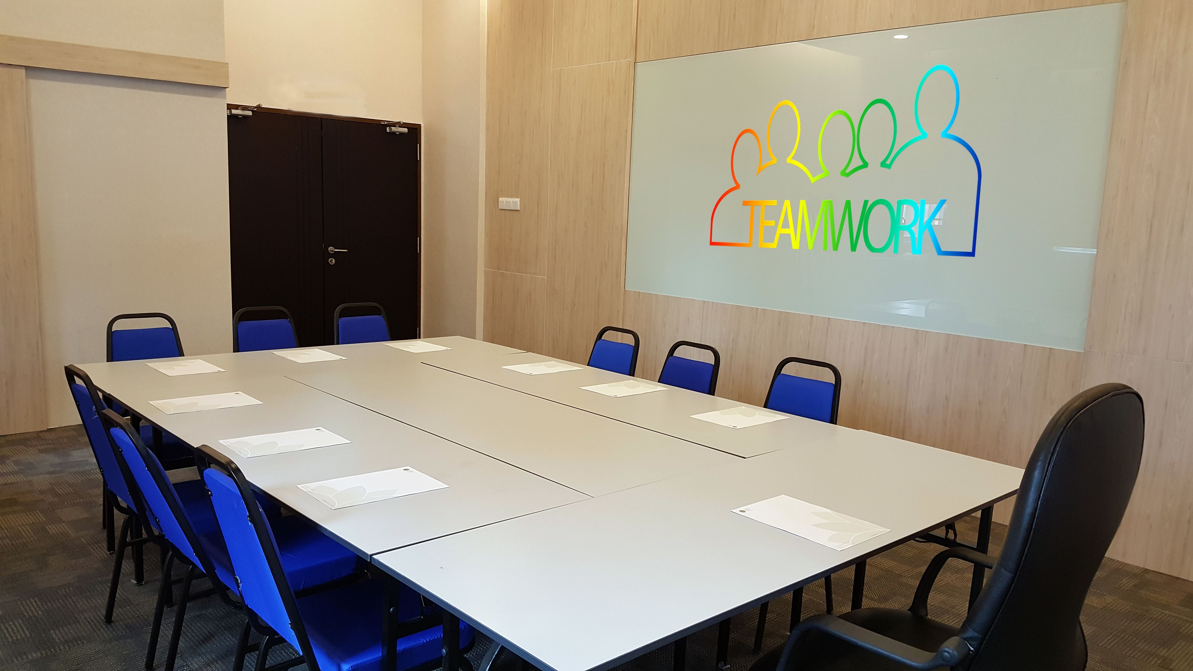Un salón de conferencias con una larga mesa, sillas azules y una pantalla mostrando 'TEAMWORK'.