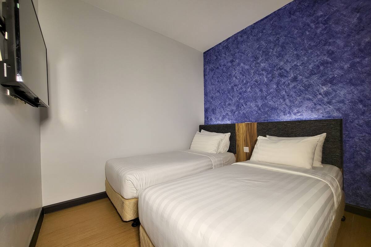 A hotel room with two beds, a wooden headboard, a blue textured wall, and a flat-screen TV mounted on the wall.