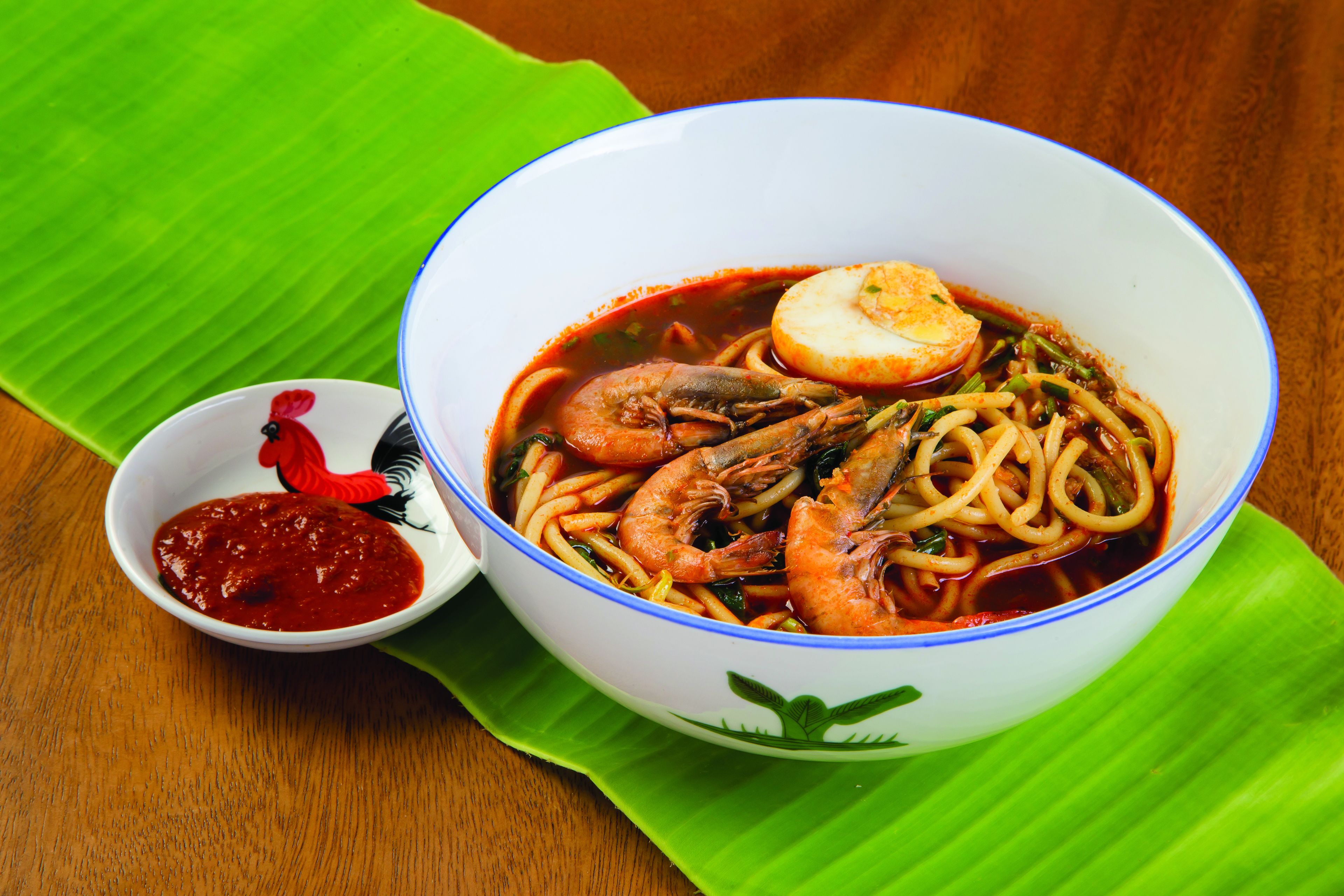 A bowl of noodle soup with shrimp and a boiled egg, served with a side of red sauce on a green leaf.