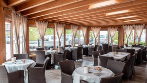 A restaurant with multiple round tables and wicker chairs, large windows with curtains, and a view of parked cars outside. - Hotel Camping Europa (Domaso)