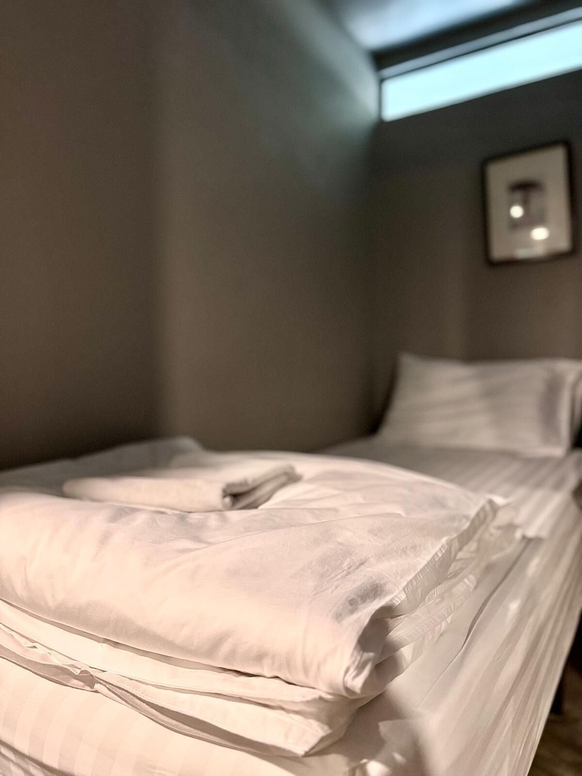 Una cama bien hecha con sábanas blancas en una habitación con pared gris y un cuadro colgado encima.