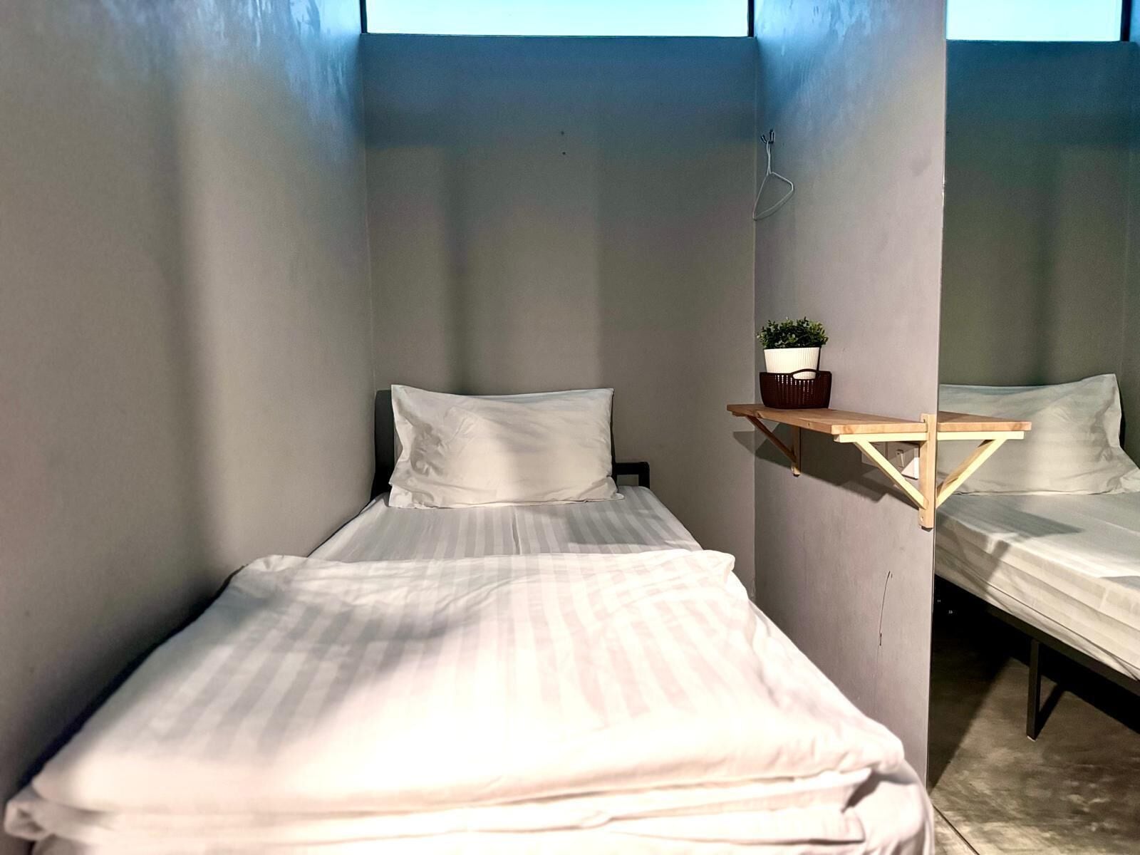 Ein kleines, gemütliches Zimmer mit zwei Einzelbetten, einem Holzregal mit einer Pflanze und einem Fenster, das natürliches Licht hereinlässt.