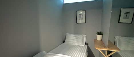 A small, modern bedroom with a single bed, a small table with a plant, and a ceiling light.