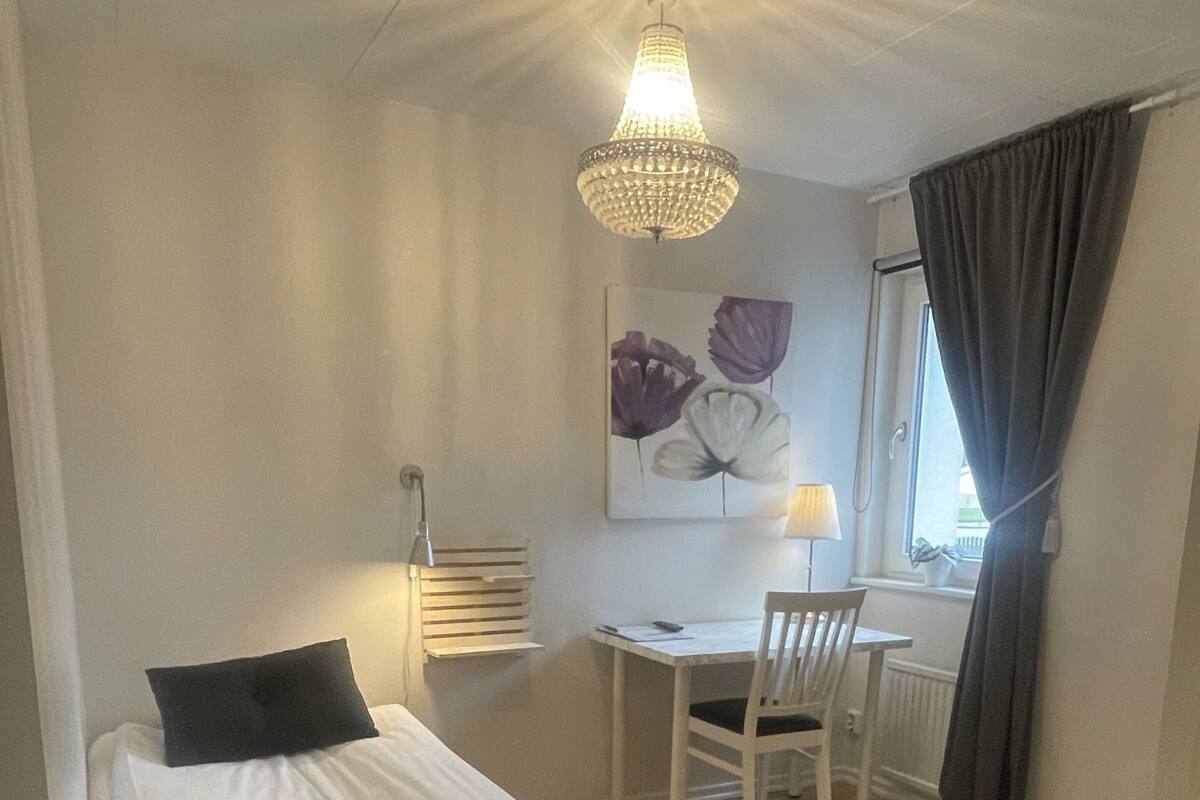 A bedroom with a bed, a desk, and a chair. There is a chandelier above the desk and a window with curtains.